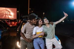 Cheerful diverse young friends smiling while taking a selfie together, standing in front of a big screen, ready to watch a movie in an open air cinema. Friendship, outdoor cinema concept