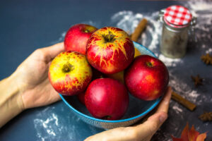 A blue bowl of red apples in women's hands for preparing a traditional American apple pie at autumn, fall. Sweet dessert for Thanksgiving. Homemade cooking, baking process. Cinnamon sticks on table.