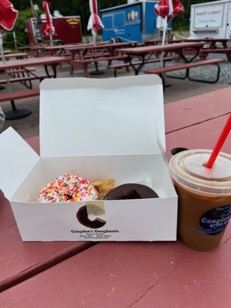Two doughnuts in a box with iced coffee on a picnic table.