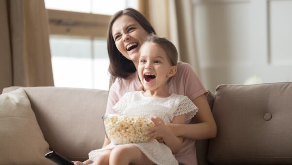 Happy young mom sit holding excited little daughter eating popcorn watch cartoon on TV together, smiling mommy or nanny have fun relaxing with small girl child enjoy movie or video with snack at home